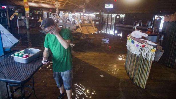 Ein Mann steht in einem überfluteten Restaurant mit Getränken und blickt auf sein Handy.