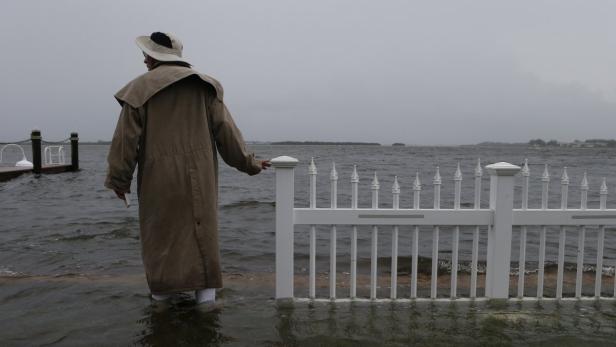 Eine Person in einem langen Mantel steht im überfluteten Wasser neben einem weißen Zaun.