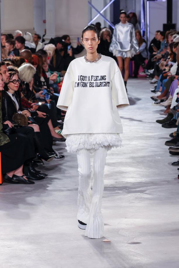 Eine Frau in einem weißen Outfit mit dem Aufdruck „I GOT A FEELING I’M GON’ BE ALRIGHT“ auf dem Laufsteg.