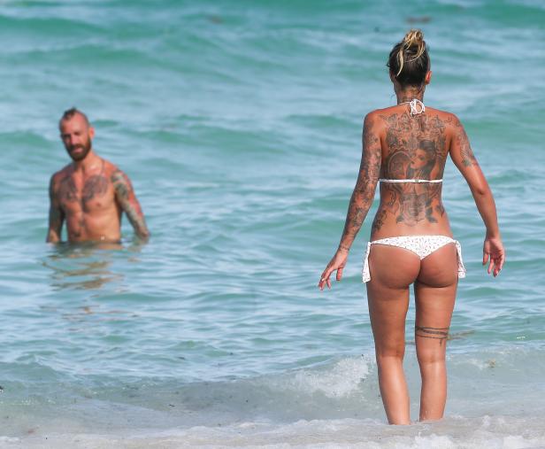 Eine Frau mit Tätowierungen steht im Meer, ein Mann im Hintergrund.