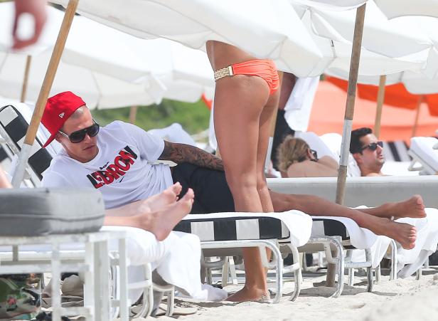 Ein Mann mit Tätowierungen und roter Kappe liegt am Strand unter einem Sonnenschirm.