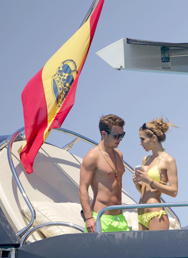 Ein Paar in Badekleidung steht auf einer Yacht unter der spanischen Flagge.