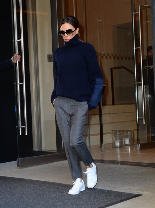 Victoria Beckham verlässt ein Gebäude in einem marineblauen Rollkragenpullover, grauer Hose und weißen Turnschuhen.