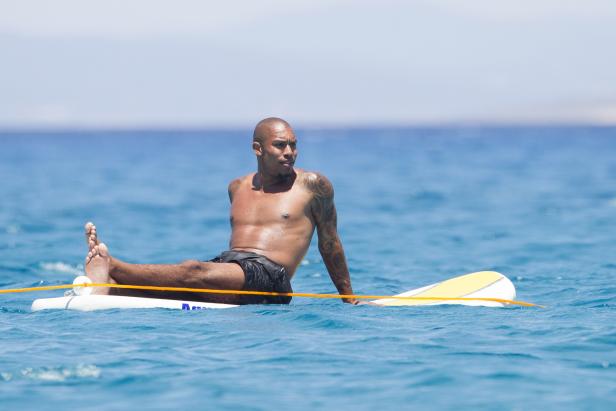 Ein tätowierter Mann sitzt auf einem Surfbrett im türkisfarbenen Wasser.