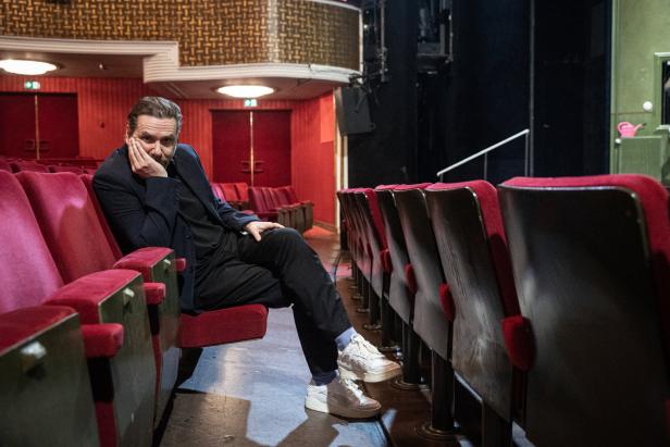 Ein Mann sitzt in einem leeren Theatersaal mit roten Sitzen.
