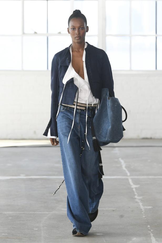 Eine Frau in Jeans, einer weißen Bluse und einer dunklen Jacke mit einer blauen Tasche.