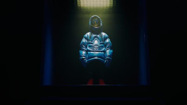 Eine Person in einer blauen Adidas-Daunenjacke mit Kapuze steht im Dunkeln.