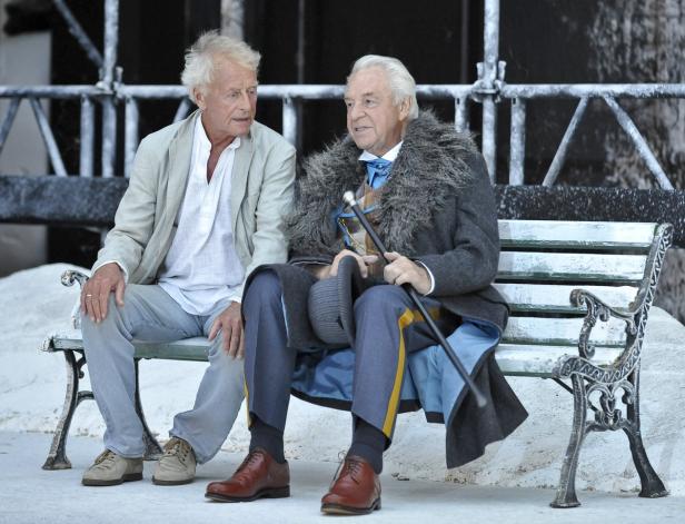 Zwei Männer sitzen auf einer Bank, der eine in legerer, der andere in formeller Kleidung mit Gehstock.