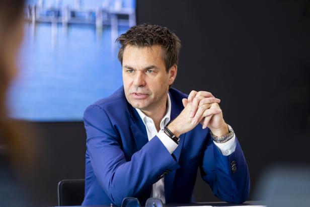 Ein Mann im blauen Anzug sitzt an einem Tisch und verschränkt nachdenklich die Hände.