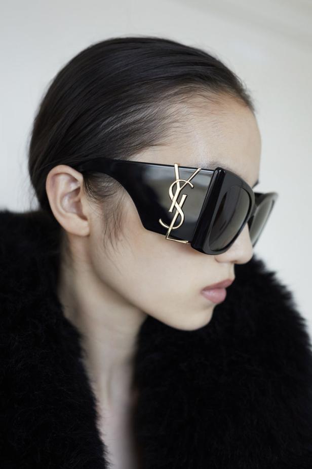 Eine Frau trägt eine schwarze Sonnenbrille mit goldenem YSL-Logo.