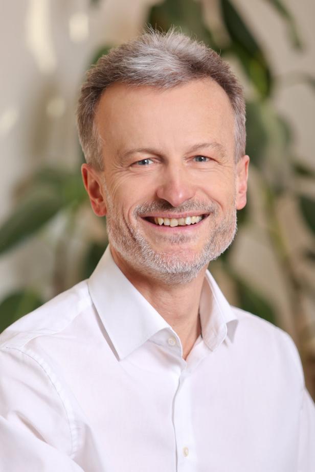 Ein lächelnder Mann mit grauen Haaren und Bart trägt ein weißes Hemd.