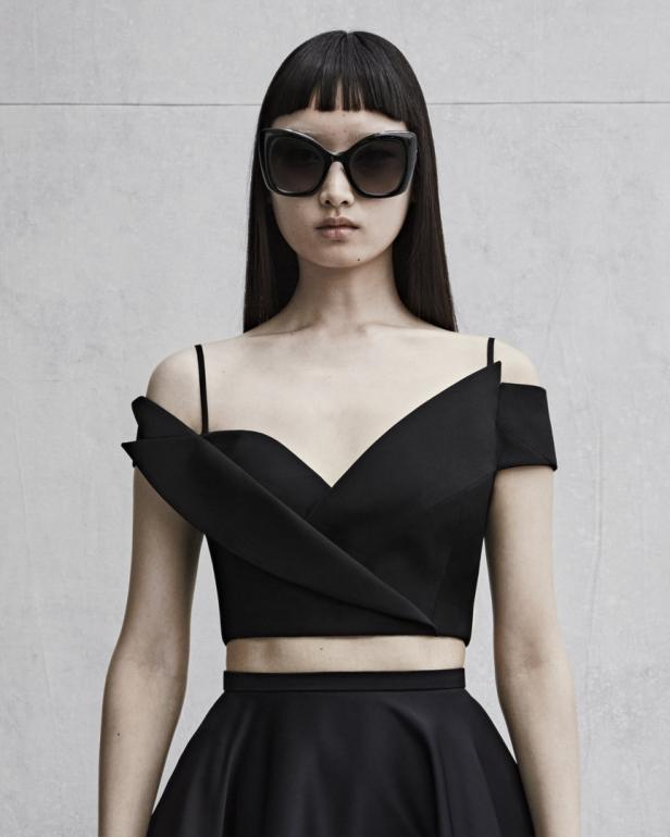 Eine Frau mit Ponyfrisur und übergroßer Sonnenbrille trägt ein schwarzes, schulterfreies Oberteil und einen Rock.