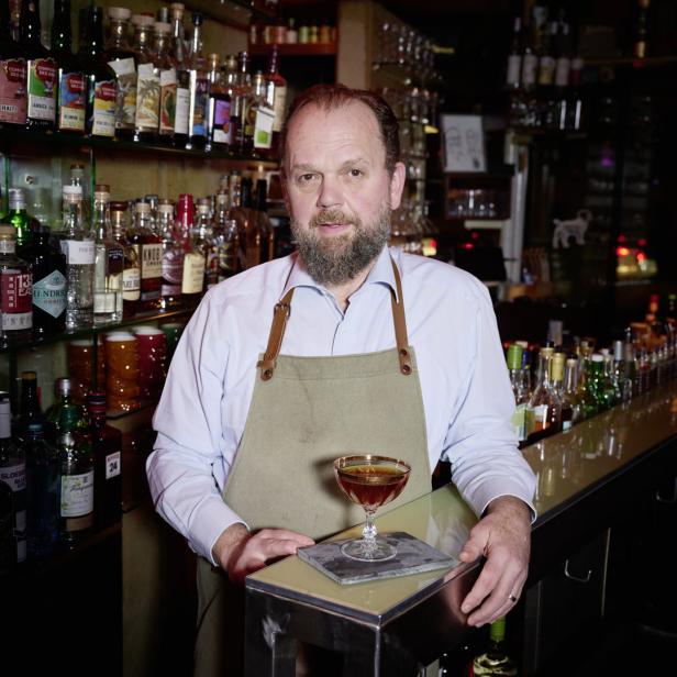 Ein Barkeeper mit Schürze steht hinter der Bar mit einem Cocktail vor sich.