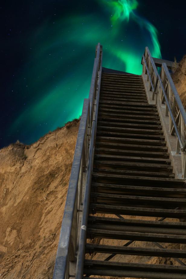 Eine lange Treppe führt zu einem Hügel unter dem grünen Polarlicht.