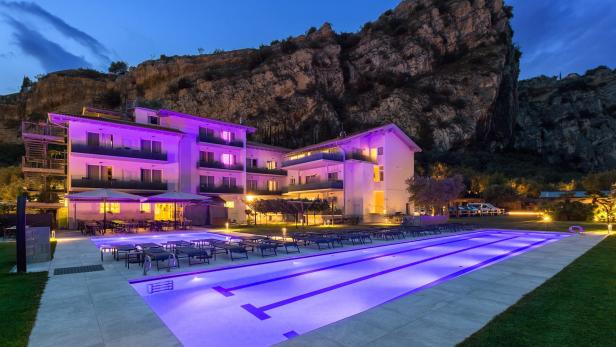 Ein Hotel mit Pool ist nachts lila beleuchtet, im Hintergrund ein Felsen.