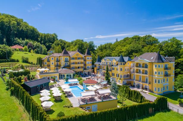 Ein gelbes Hotelresort mit Pool und gepflegten Gärten vor einem bewaldeten Hügel.