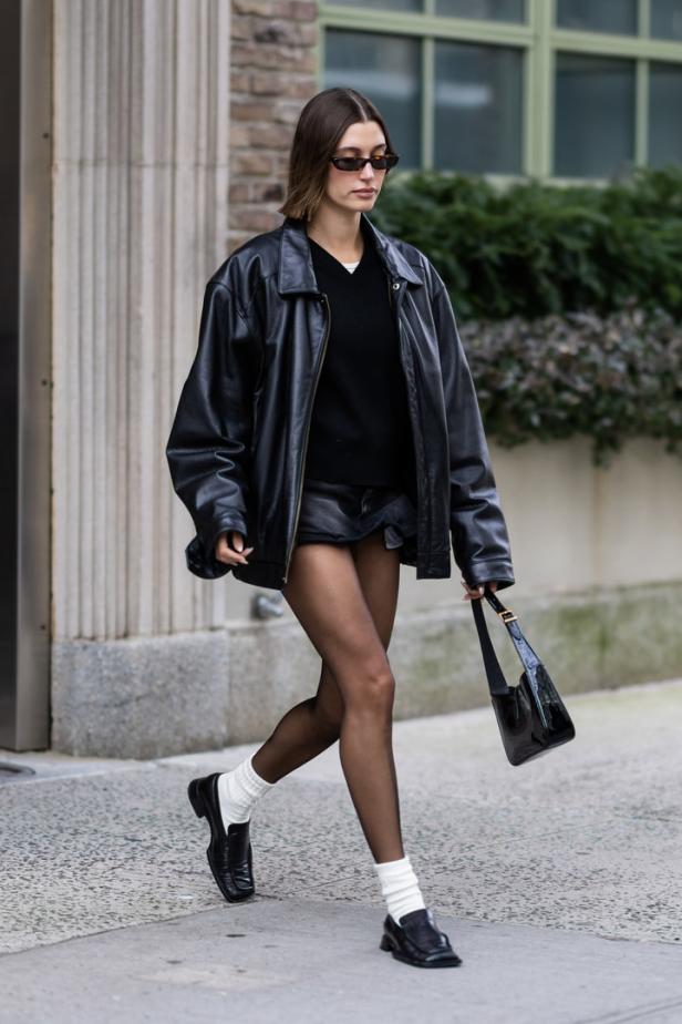 Eine Frau in Lederjacke, Minirock und Loafern mit Socken geht die Straße entlang.