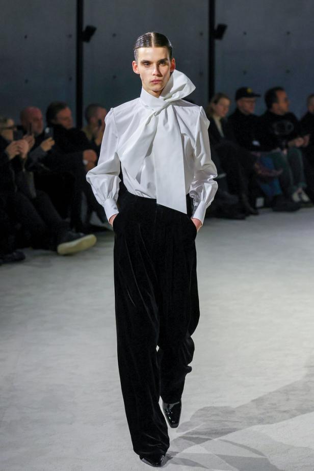Ein Model mit weißer Bluse mit Schleife und schwarzer Hose auf einem Laufsteg.