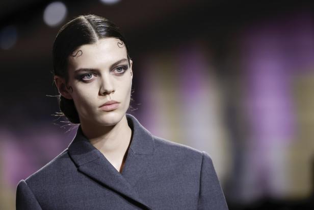 Eine Frau mit dunklem Haar und auffälligem Make-up trägt einen grauen Blazer.