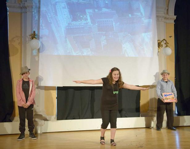 Eine junge Frau steht mit ausgestreckten Armen auf einer Bühne, flankiert von zwei Jungen.