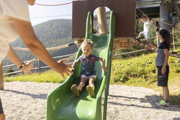 Ein Kind rutscht fröhlich eine grüne Rutsche auf einem Spielplatz hinunter.