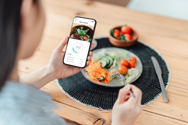 Eine junge Frau isst einen Salat und benutzt eine Kalorienzähler-App auf ihrem Smartphone, um ihre tägliche Kalorienzufuhr zu überwachen. Sie erstellt einen Diätplan, um Gewicht zu verlieren.