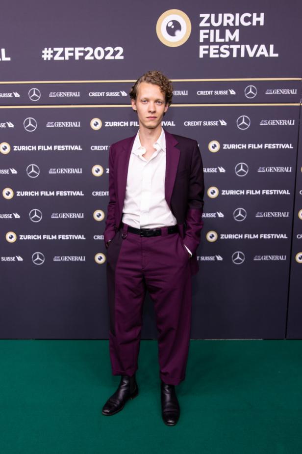 Ein Mann im Anzug posiert vor einer Wand des Zurich Film Festivals 2022.