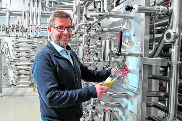 Ein Mann mit Brille hält ein Glas Bier vor einer Anlage mit Rohren.