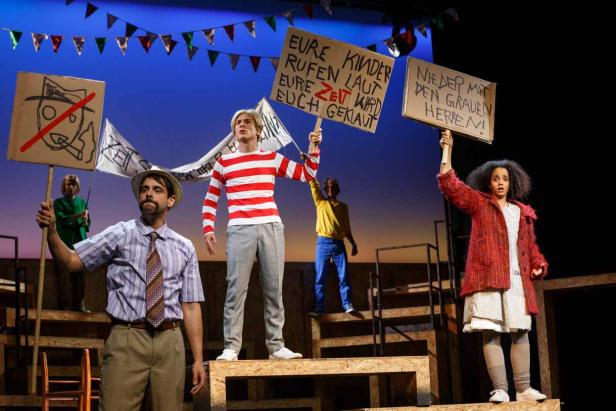 Eine Theaterszene mit Schauspielern, die Schilder mit Aufschriften halten.