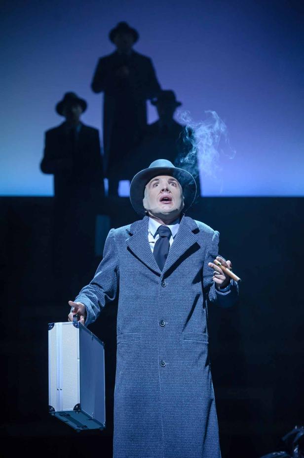 Ein Mann im Anzug und Hut mit Zigarre und Koffer auf einer Bühne.