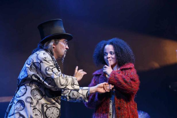 Ein Mann mit Zylinder und eine junge Frau mit lockigem Haar stehen auf einer Bühne. Die Frau hält einen Finger an den Mund.