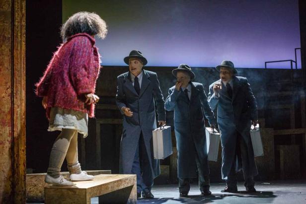 Eine junge Frau steht vor drei Männern in Anzügen und Hüten auf einer Bühne.