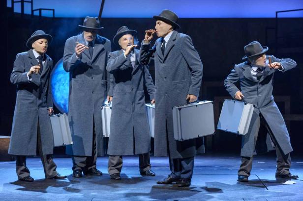 Fünf Personen in grauen Mänteln und Hüten stehen mit Zigarren und Aktenkoffern auf einer Bühne.