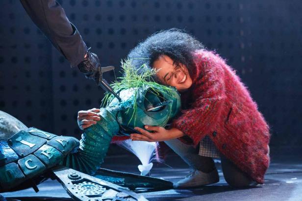 Eine Frau umarmt eine grüne Schildkrötenpuppe, während eine Hand mit einem Messer droht.