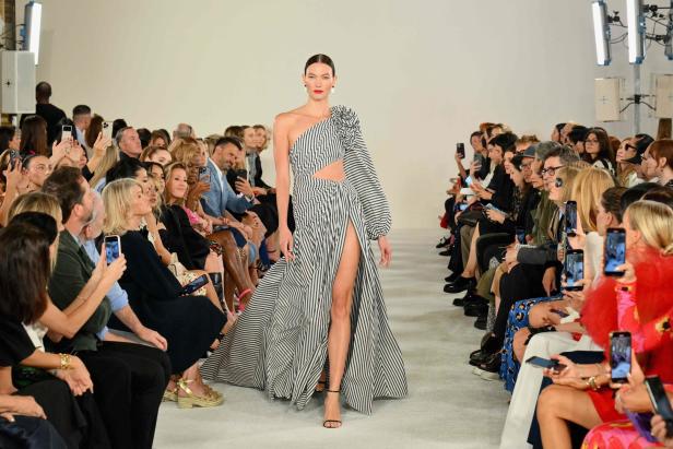 Ein Model präsentiert ein gestreiftes, asymmetrisches Kleid auf dem Laufsteg vor einem Publikum.