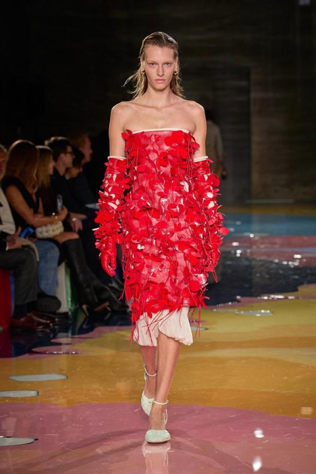 Ein Model präsentiert ein rotes, trägerloses Kleid mit Blütenapplikationen und passende Handschuhe auf dem Laufsteg.