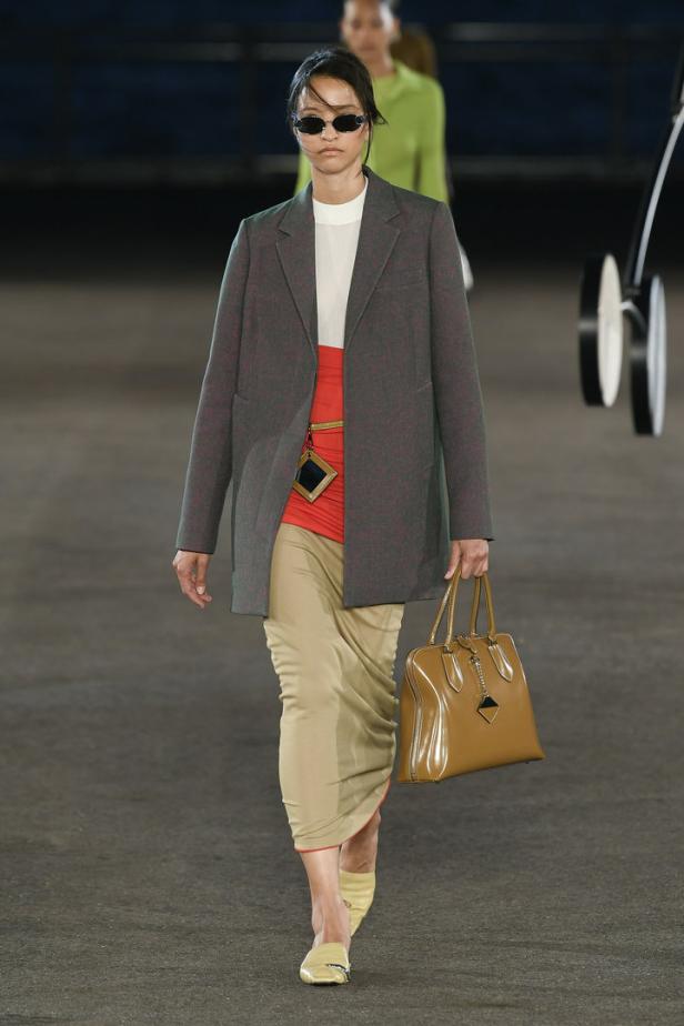 Eine Frau in Blazer, Rock und Sonnenbrille läuft auf einem Laufsteg mit einer Handtasche.