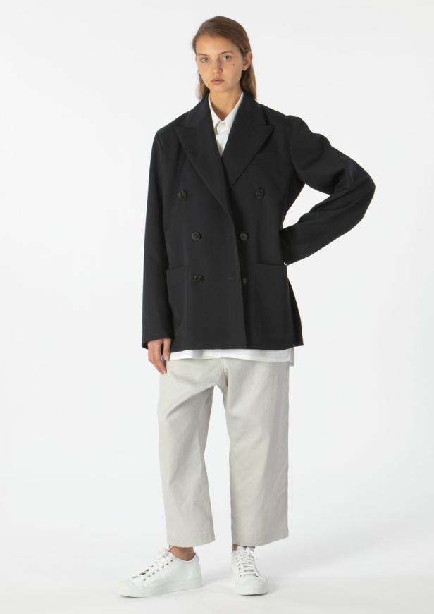 Eine Frau in einem übergroßen schwarzen Blazer, hellen Hosen und weißen Turnschuhen vor einem weißen Hintergrund.