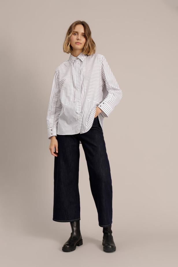 Eine Frau mit blonder Bob-Frisur trägt eine gestreifte Bluse, dunkle Jeans und schwarze Stiefel.