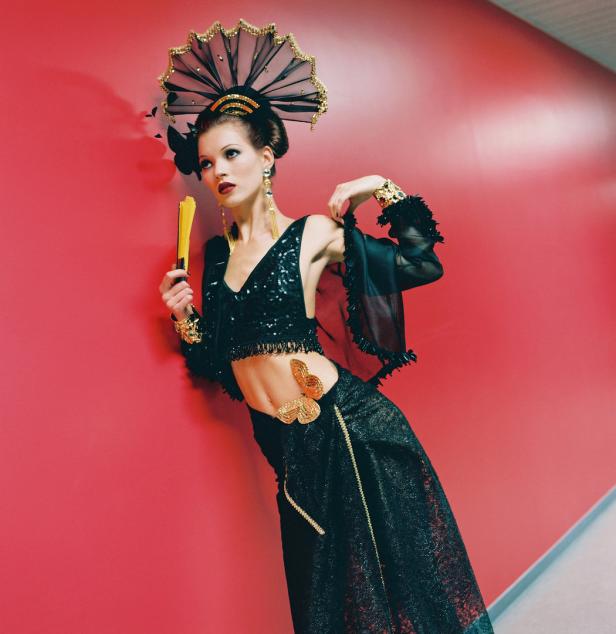Eine Frau in einem schwarzen, bauchfreien Outfit mit goldenen Accessoires posiert vor einer roten Wand.