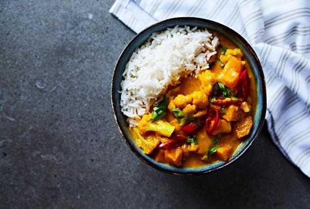 Eine Schüssel mit Curry, Reis, Blumenkohl und roten Paprika steht auf einem Tisch.