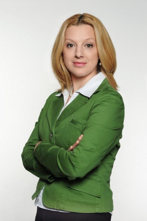 Eine Frau mit blonden Haaren und grünem Blazer steht mit verschränkten Armen da.