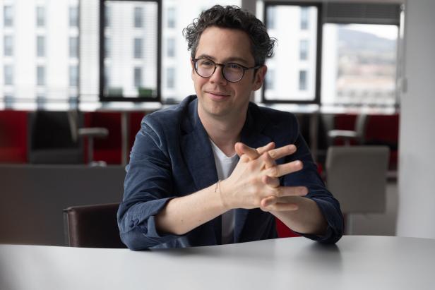 Ein Mann mit Brille und lockigen Haaren sitzt lächelnd an einem Tisch in einem modernen Büro.