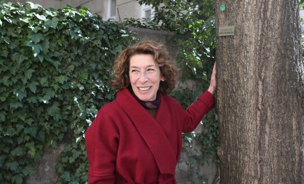 Eine lächelnde Adele Neuhauser in einem roten Mantel lehnt an einem Baum.