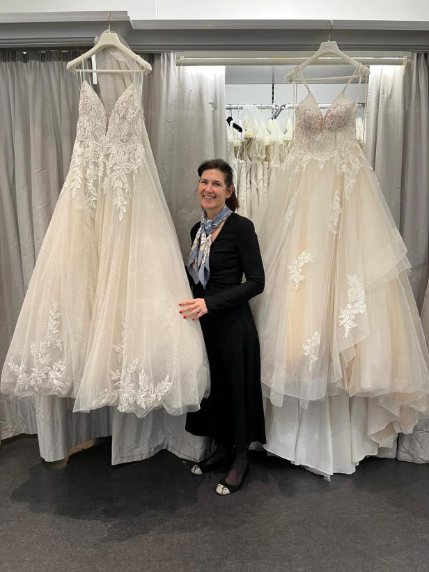 Eine Frau steht in einem Brautmodengeschäft zwischen zwei Hochzeitskleidern.