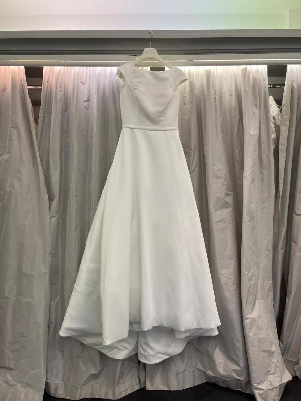 Ein schlichtes, weißes Brautkleid hängt an einem Kleiderbügel vor einem Vorhang.