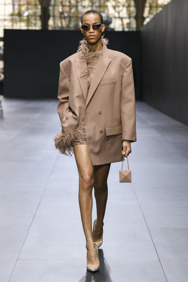 Eine Frau in einem übergroßen Blazer mit Federn und einer kleinen Handtasche auf dem Laufsteg.