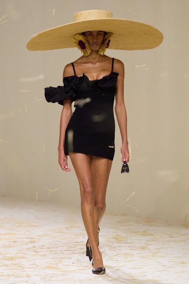 Eine Frau mit einem schwarzen Kleid und einem übergroßen Strohhut auf dem Laufsteg.