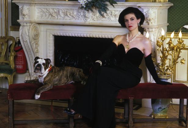 Eine elegant gekleidete Frau sitzt mit einem Bulldog vor einem Kamin.