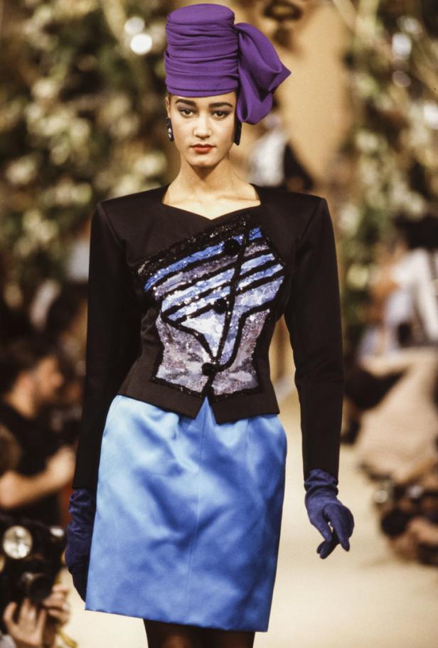 Eine Frau in einem schwarzen Blazer mit Pailletten und einem blauen Rock mit passendem Turban auf dem Laufsteg.